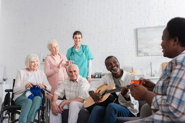 老人ホームで看護師の近くにアフリカ系アメリカ人の友人を見て糸とアコースティックギターを持つ陽気な異人種間の高齢者 — ストック写真