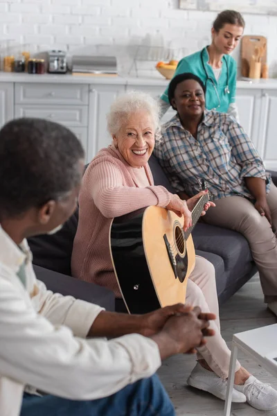 Smiling elderly woman playing acoustic guitar near multiethnic friends and blurred nurse in nursing home