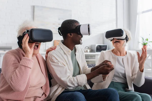 Excited interracial people gaming in vr headsets on couch in nursing home