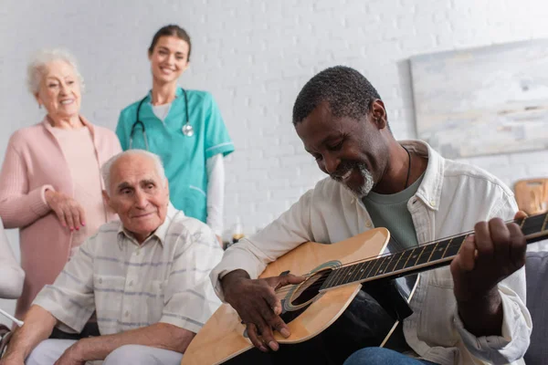 Pensionista Afroamericano Tocando Guitarra Acústica Cerca Amigos Enfermeras Residencia Ancianos — Foto de Stock