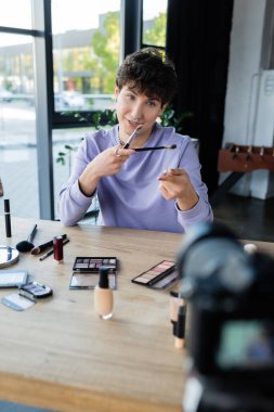 Transgender blogger holding cosmetic brushes near digital camera in studio  clipart