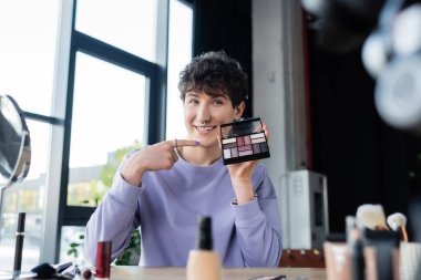 Smiling transgender makeup artist pointing at eye shadows  clipart