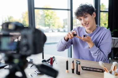 Smiling transgender person holding cosmetics near digital camera  clipart