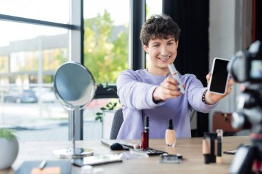 Smiling transgender person holding mascara and smartphone near decorative cosmetics and digital camera  clipart