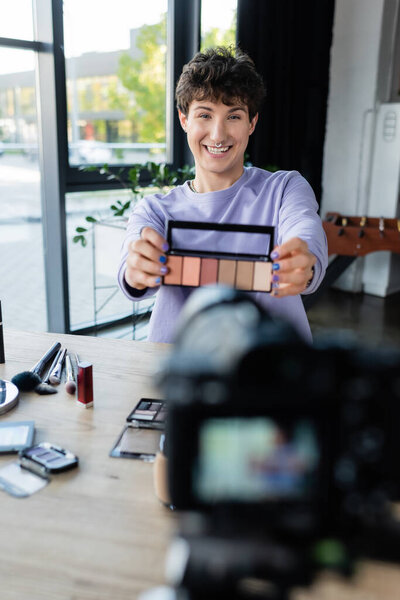 Smiling transgender makeup artist showing eye shadows at digital camera 