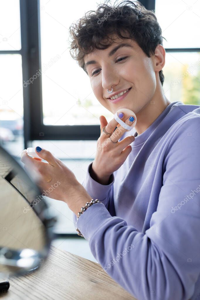 Smiling transgender person applying face powder near blurred mirror 
