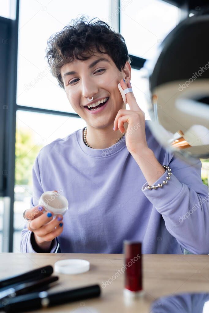 Happy transgender person applying face powder near blurred decorative cosmetics and mirror 