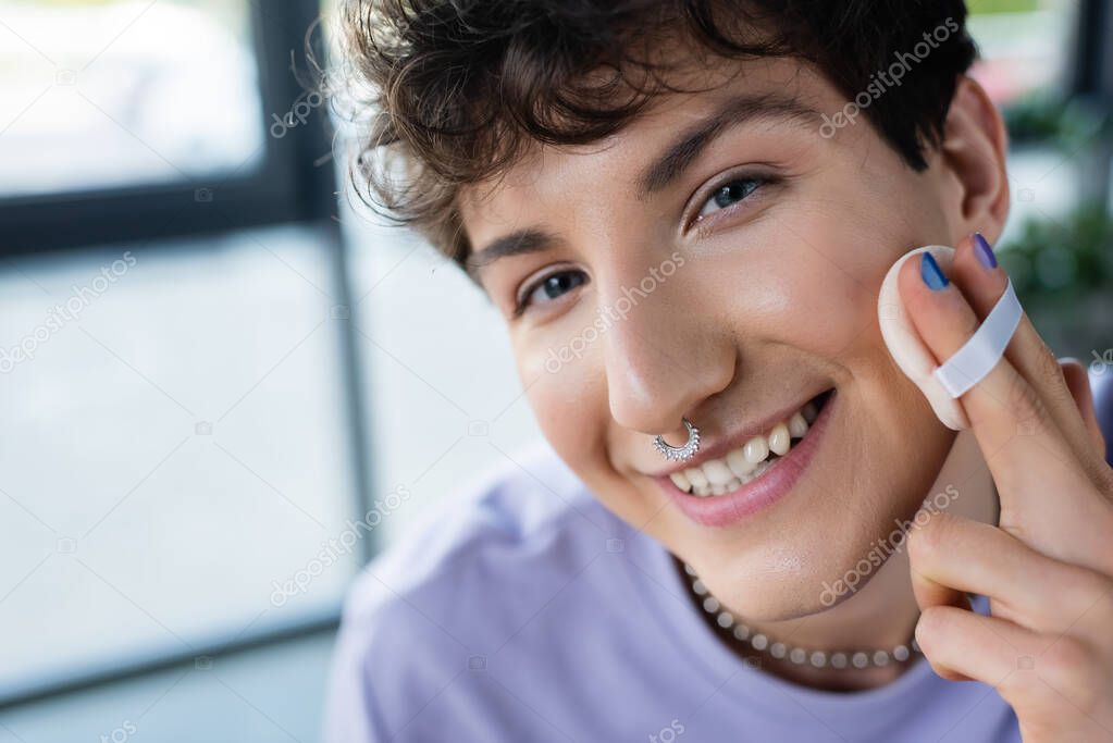 Portrait of smiling transgender person with piercing applying face powder and looking at camera 