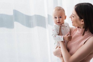 happy brunette woman holding baby boy near white curtain at home clipart