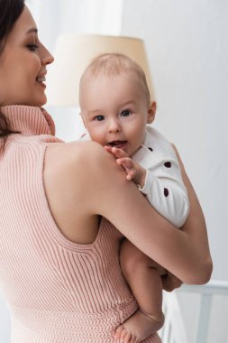 Küçük çocuk evdeki annesinin elinde kameraya bakıyor.