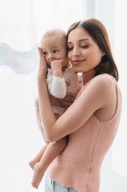 Gözleri kapalı neşeli bir kadın. Evinde yeni yürümeye başlayan oğlunu kucaklıyor.