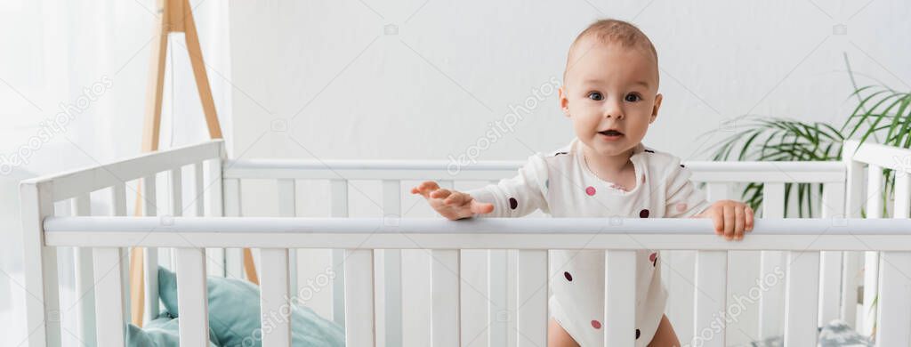 baby in romper looking at camera while standing in crib, banner