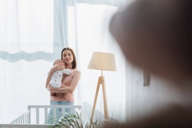 Gözleri kapalı neşeli bir kadın oğlunu beşiğin yanında tutuyor, ön planda bulanıklık var.