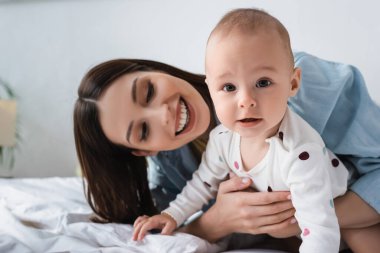 Küçük oğlunu kucaklayan gülümseyen bir kadın yatakta kameraya bakıyor.
