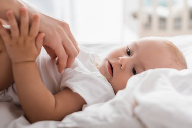 Küçük çocuk beyaz bir yatakta yatarken annesinin eline dokunuyor.