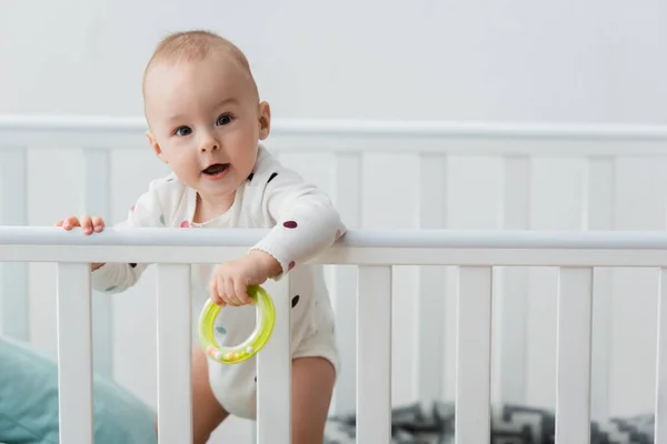 ベビーベッドに立ってカメラを見ながらガラガラリングを持っている男の子 — ストック写真