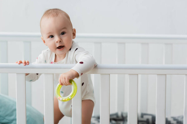 little boy holding rattle ring while standing in crib and looking at camera