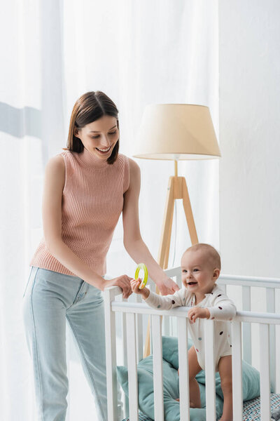 brunette woman smiling near little son standing in crib with rattle ring