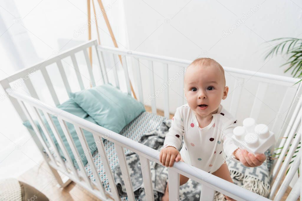 high angle view of little child holding toy building block while standing in crib