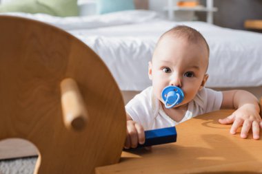 Emzik taşıyan bebek oyuncak yapı taşı tutarken sallanan atın yanındaki kameraya bakıyor.