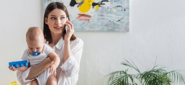 Gülümseyen kadın küçük oğlunu kucağında tutarken cep telefonu ve afiş ile konuşuyor.