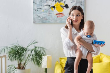 Evde yeni yürümeye başlayan çocukla çalışan bir kadın cep telefonuyla konuşuyor.