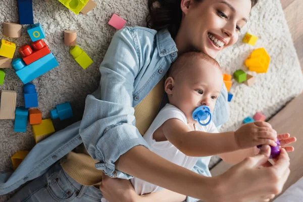 Küçük oğlu çok renkli blokların yanında yerde yatan neşeli kadının yüksek açılı görüntüsü.