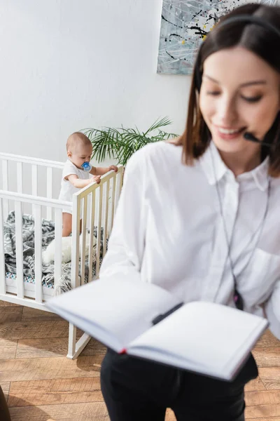 Blurred Woman Headset Holding Empty Notebook While Working Son Crib — Stock Photo, Image