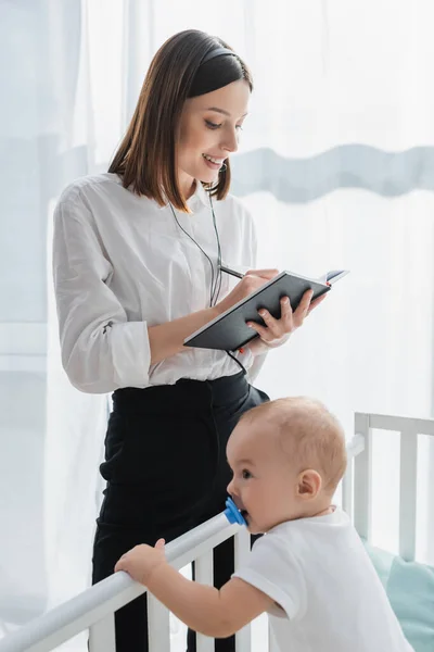 Frau Mit Headset Schreibt Notizbuch Neben Baby Kinderbett — Stockfoto