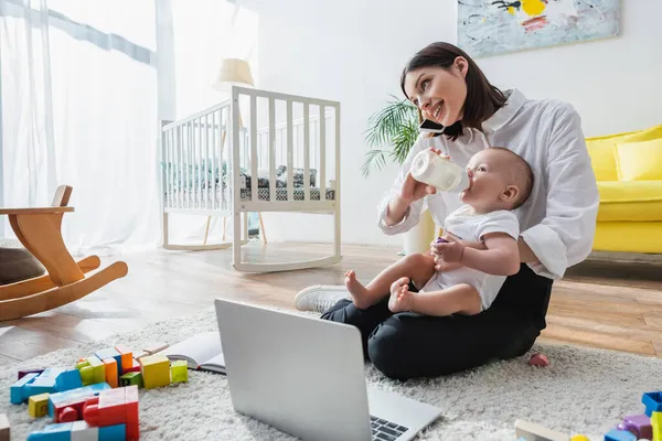 Smiling Woman Talking Smartphone While Feeding Son Floor Laptop Building — Stock Photo, Image