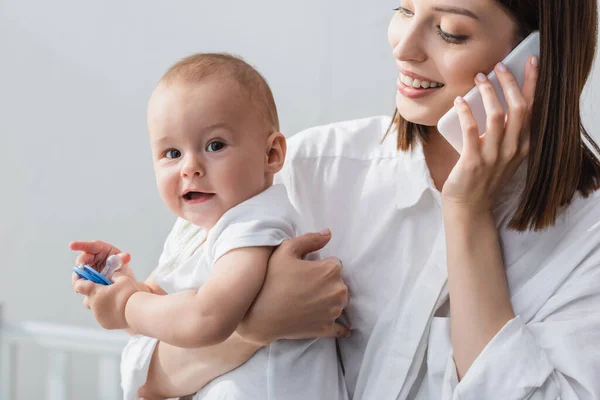 Smilende Kvinne Som Snakker Mobiltelefon Mens Hun Holder Småbarn Hjemme – stockfoto