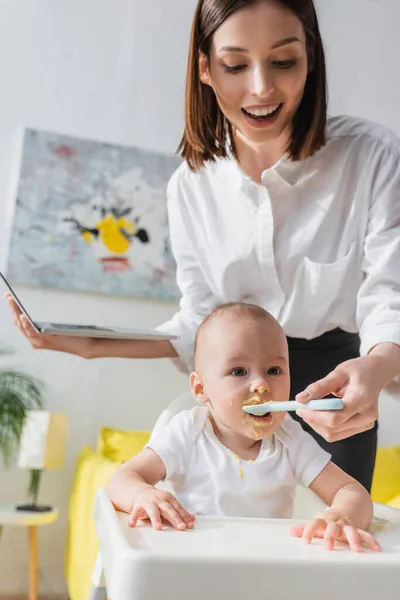 Leende Kvinna Hålla Laptop Medan Mata Barnet Pojke Med Puré — Stockfoto