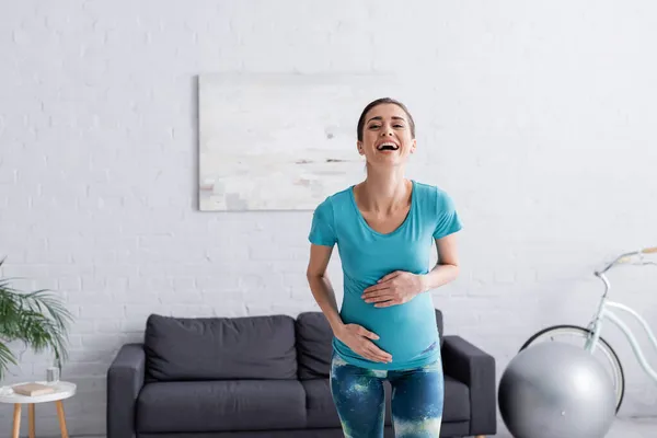 元気な妊婦さんがお腹に触れてリビングルームで笑って — ストック写真
