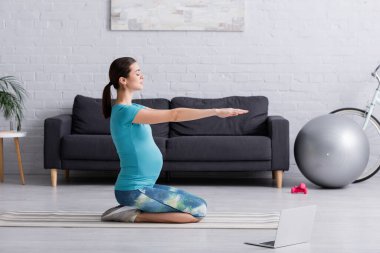 Spor kıyafetli hamile bir kadının spor salonunda spor minderinde dizüstü bilgisayarın yanında çalışmasının yan görüntüsü.