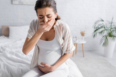 young pregnant woman feeling nausea and covering mouth in bedroom  clipart