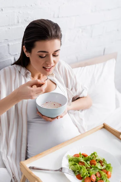 Feliz Mujer Embarazada Comiendo Avena Cerca Bandeja Con Ensalada — Foto de Stock