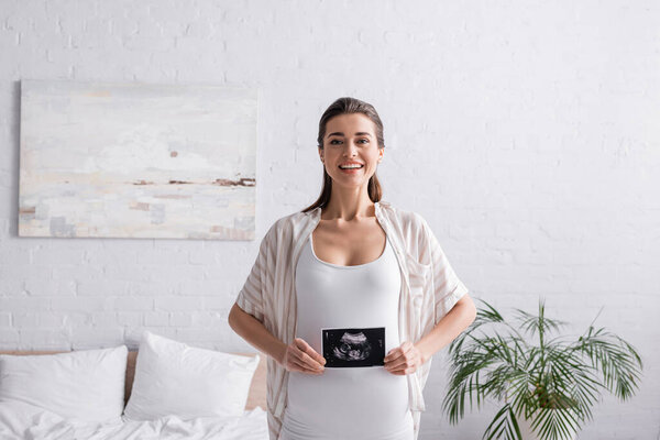smiling and pregnant woman holding ultrasound scan in bedroom 