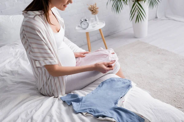 ベッドルームで赤ちゃんの服を持って幸せな妊婦のクロップドビュー — ストック写真