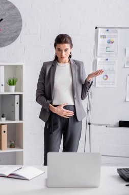 young pregnant businesswoman gesturing near flip chart with chats and graphs during video call on laptop clipart