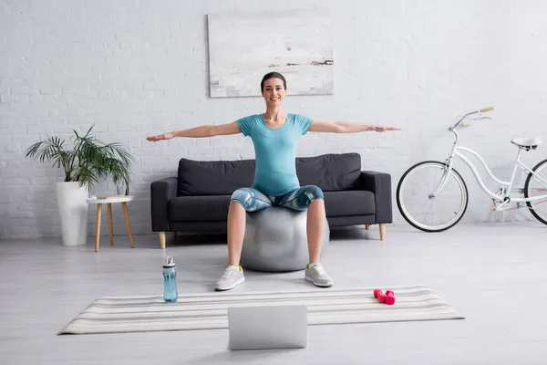快乐的孕妇穿着运动服在客厅的笔记本电脑旁的健身球上锻炼 — 图库照片