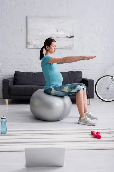 孕妇穿着运动服在客厅里笔记本电脑旁边的健身球上运动 — 图库照片