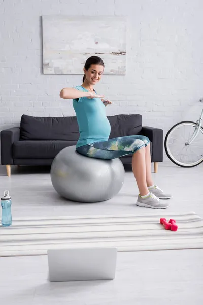 孕妇穿着运动服在客厅里笔记本电脑旁的健身球上运动 — 图库照片