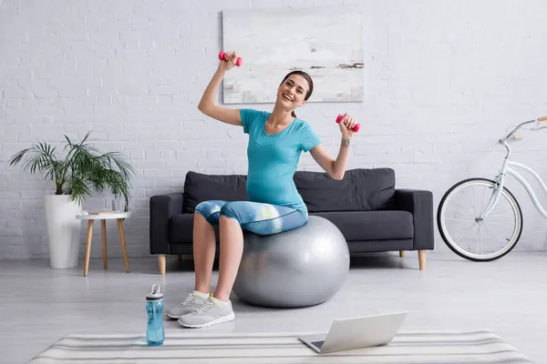 快乐的孕妇带着哑铃在笔记本电脑旁的健身球上锻炼 — 图库照片
