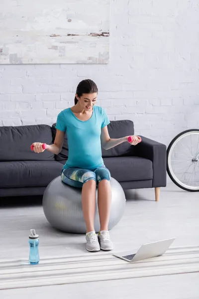 快乐的怀孕女运动员在运动瓶子和笔记本电脑旁边用哑铃进行健身球运动 — 图库照片