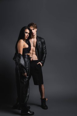 Deri ceketli, kaslı bir adam. Gri elbiseli esmer kadının yanında duruyor.