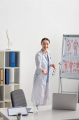 usmívající se lékař ukazuje rukou při videohovoru, zatímco stojí v blízkosti flip chart s anatomickými obrázky