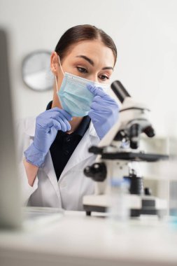Tıbbi maskeli bilim adamı laboratuvarda dizüstü bilgisayarın yanında mikroskoba bakıyor. 