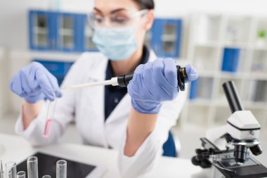 Tıbbi maskeli bulanık bilim adamı test tüpleri, mikroskop ve dijital tabletin yanında elektronik pipet tutuyor. 