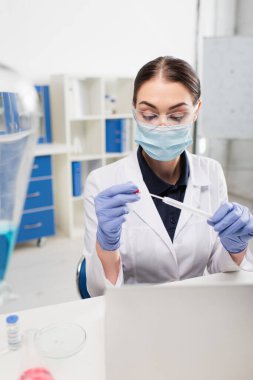 Gözlüklü ve lateks eldivenli bilim adamı laboratuvarda dizüstü bilgisayarın yanında test yapıyor. 