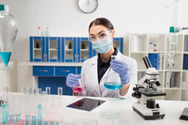 Bilim adamı dijital tabletin yanında boş ekranlı mataralar ve laboratuarda mikroskop tutuyor. 